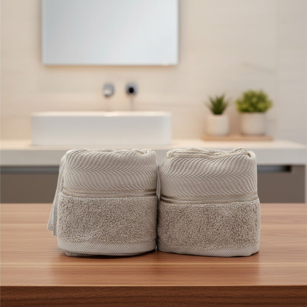 Two beige Cotton Bath & Face Towel Set on a wooden surface with a blurred bathroom background