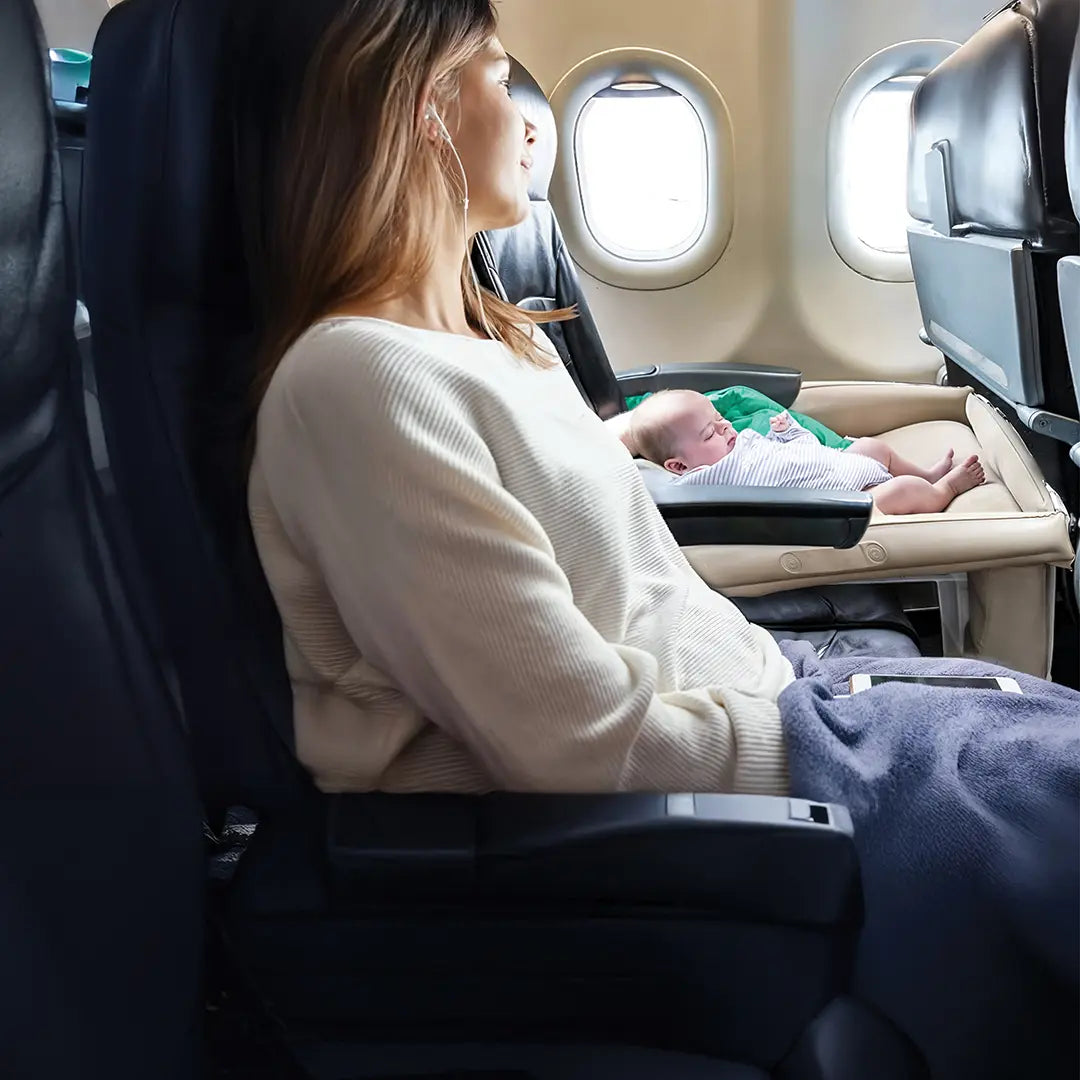 A woman sitting in an airplane seat with a baby sleeping at the Green Lion Inflatable Toddler Travel Bed.
