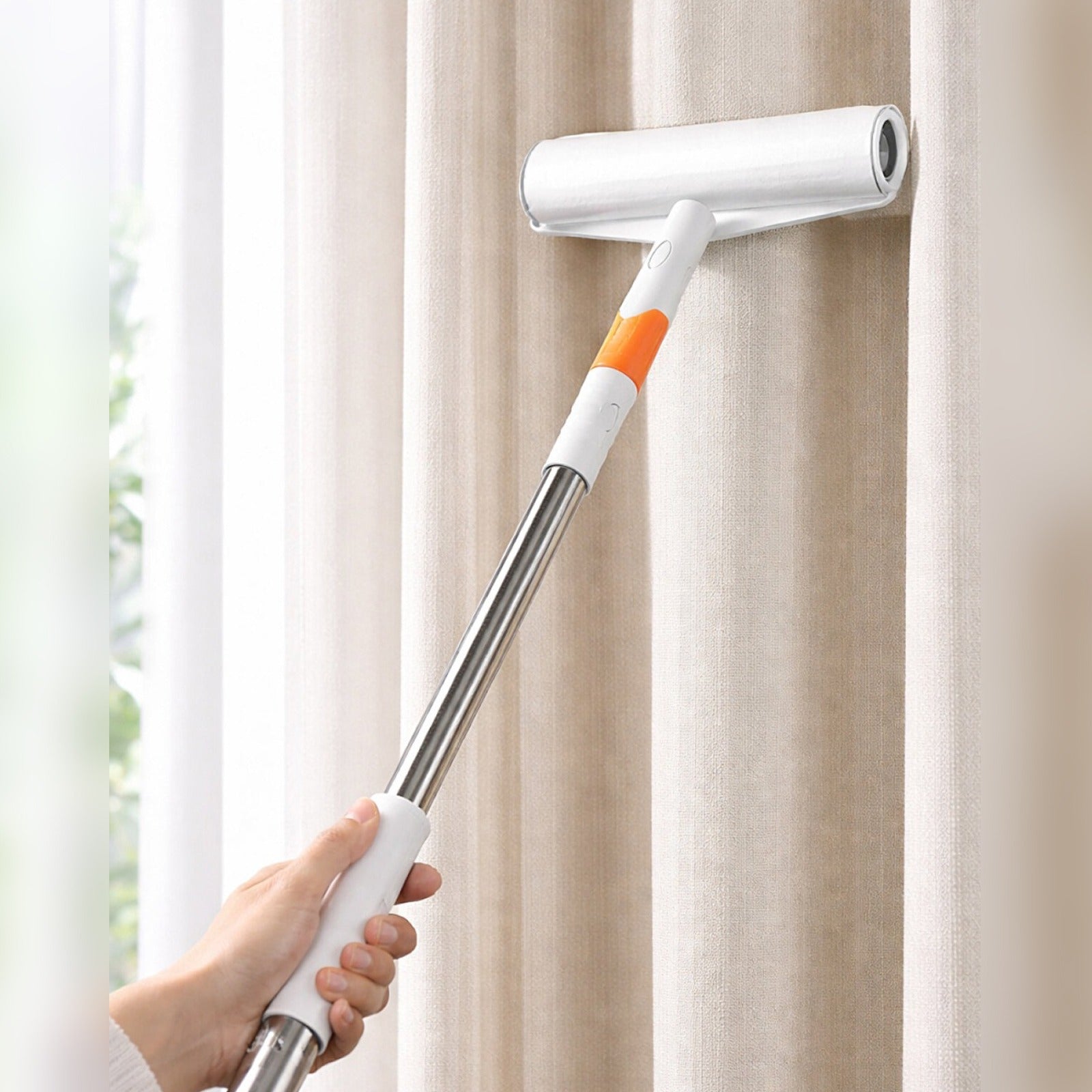 A hand holds a Sticky Mop against a beige curtain, showing the white and orange roller attachment in use.